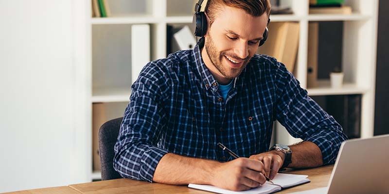 lächelnder Mann mit Kopfhörern, der Notizen macht, während er an einem Online Seminar teilnimmt